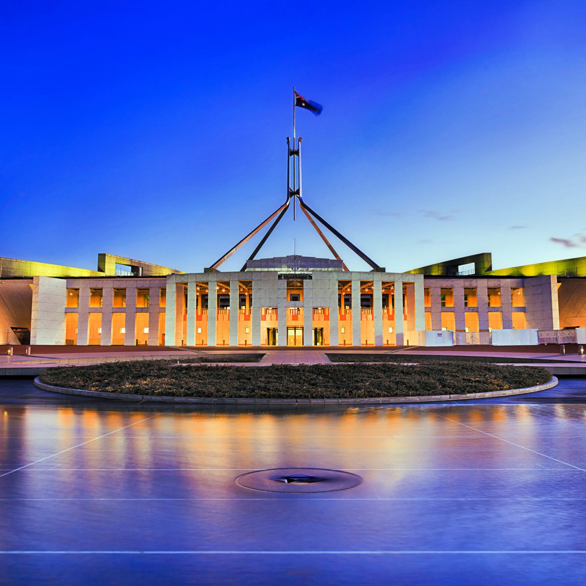 Facade of new parliament house in Canberra on capitol hill at sunset with bright illumination reflecting in blurred waters of pool.